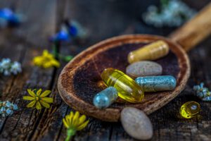 Tabletten und Kapseln in verschiedenen Farben auf einem Holzlöffel umgeben von Blumen