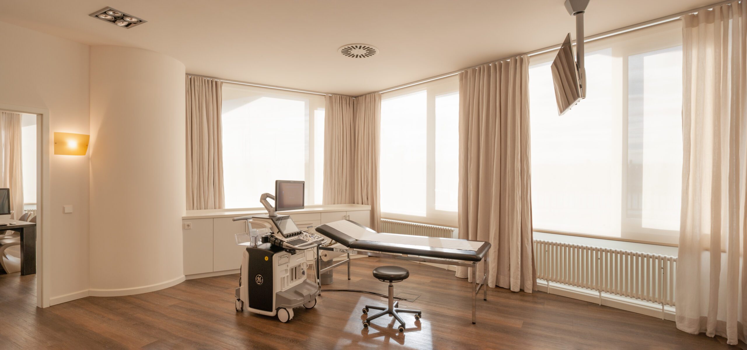 Heller Untersuchungsraum in der mamonova Praxis mit moderner Ultraschalltechnik, Untersuchungsliege und großen Fenstern mit Tageslicht.