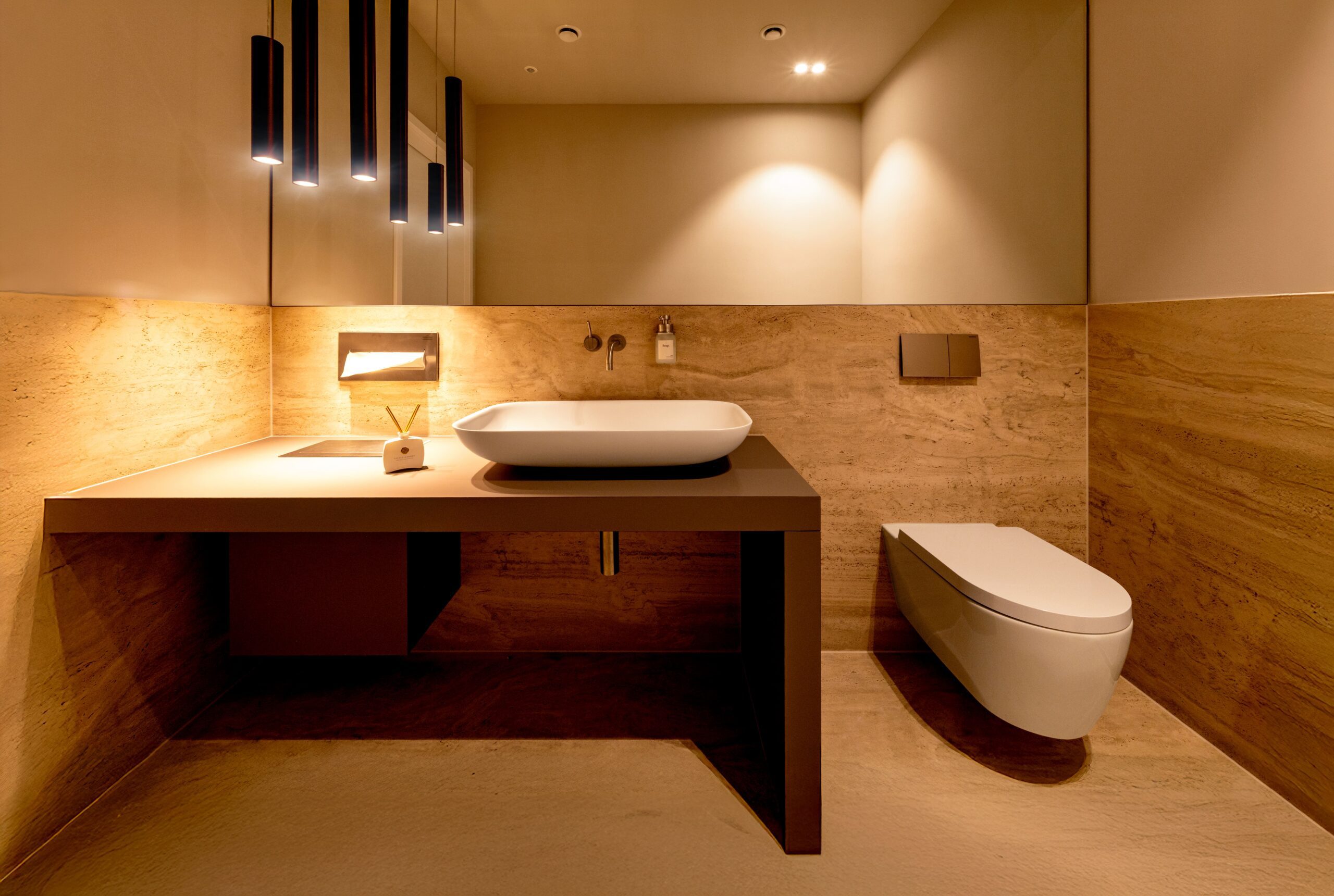 Modern gestaltetes Badezimmer der mamonova Praxis in Lindenthal mit heller Natursteinoptik, Designerwaschbecken, wandhängender Toilette und stimmungsvoller Beleuchtung.