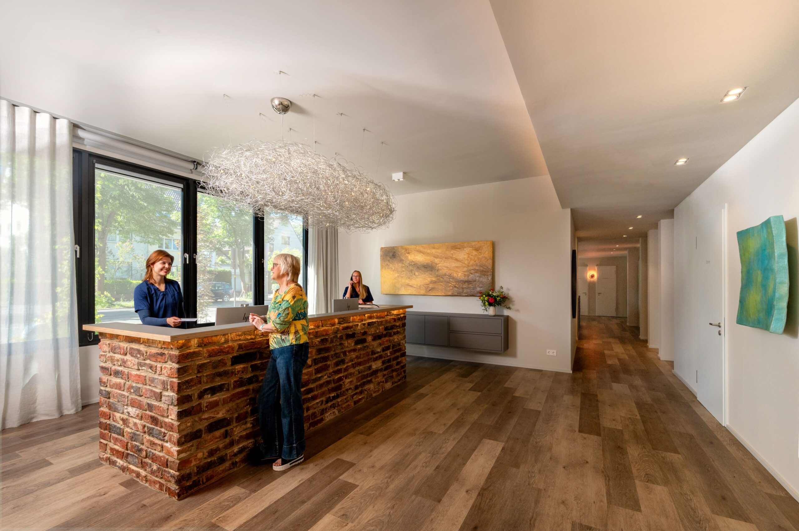 Empfangsbereich der mamonova Praxis in Lindenthal mit großen Fenstern, moderner Einrichtung und freundlicher Begrüßung einer Patientin durch eine Mitarbeiterin am Tresen. Im Hintergrund telefoniert eine weitere Mitarbeiterin.