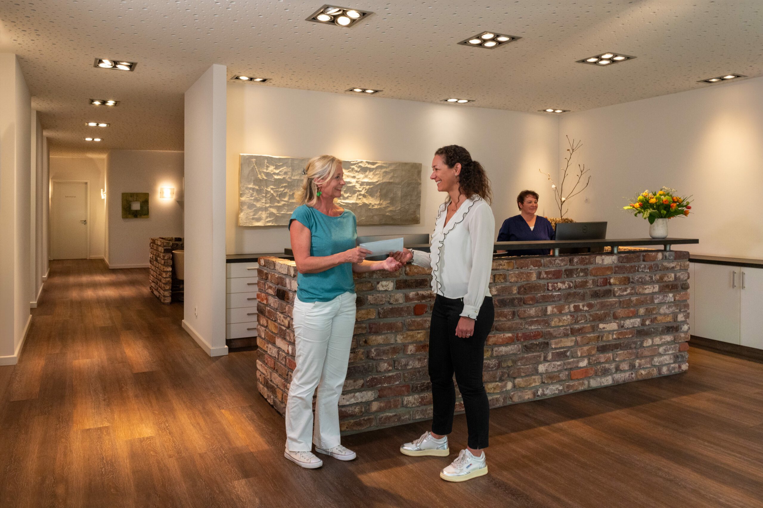 Freundliche Übergabe von Unterlagen zwischen Ärztin und Patientin vor dem Empfang der mamonova Praxis. Im Hintergrund arbeiten Mitarbeitende am Tresen in heller, moderner Umgebung.