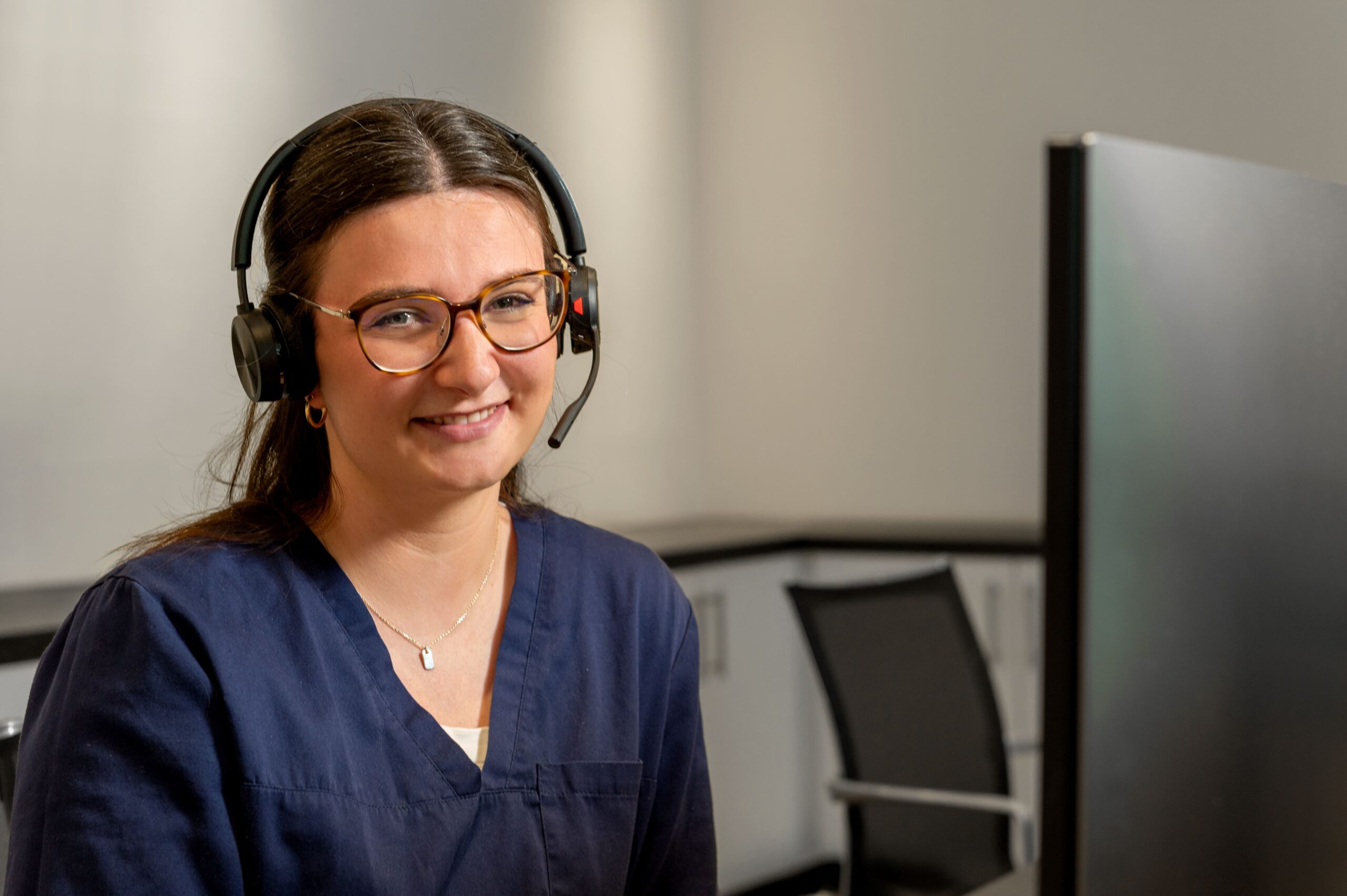 Mitarbeiterin der mamonova Praxis mit Headset am Empfangsplatz. Sie lächelt freundlich in die Kamera, im Hintergrund ist ein Arbeitsplatz mit Monitor sichtbar.