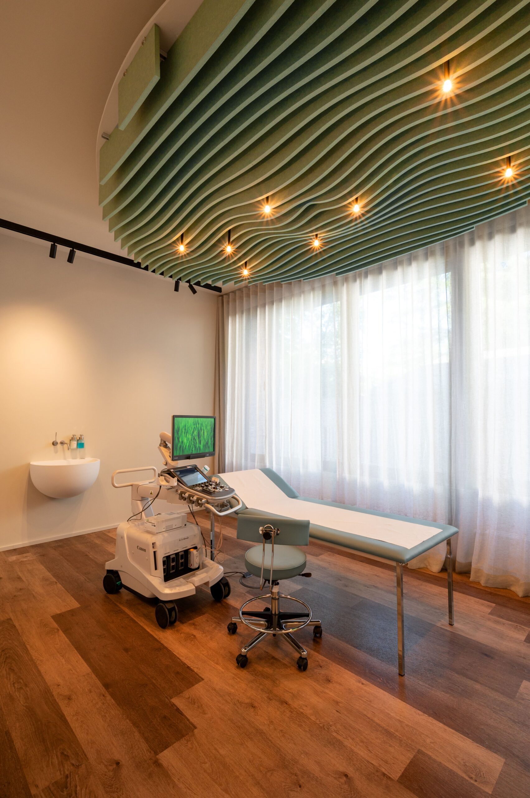Modernes Untersuchungszimmer der mamonova Praxis in Lindenthal mit Ultraschallgerät, Untersuchungsliege und beruhigend gestaltetem Deckenelement in organischer Form.