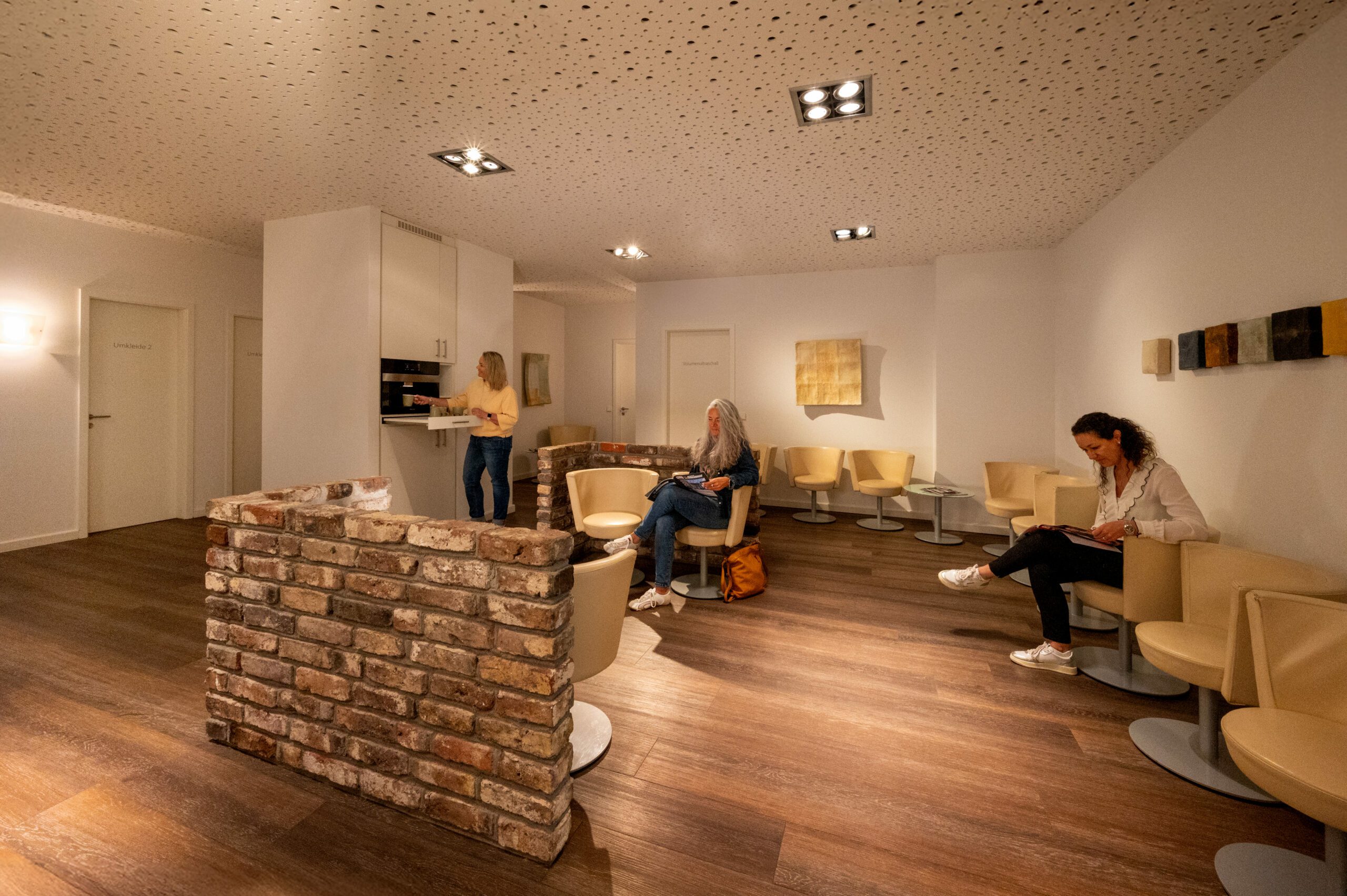 Wartebereich der mamonova Praxis mit warmem Licht, modernen Sitzgelegenheiten und entspannter Atmosphäre. Zwei Patientinnen sitzen lesend, eine weitere bedient die Kaffeestation.