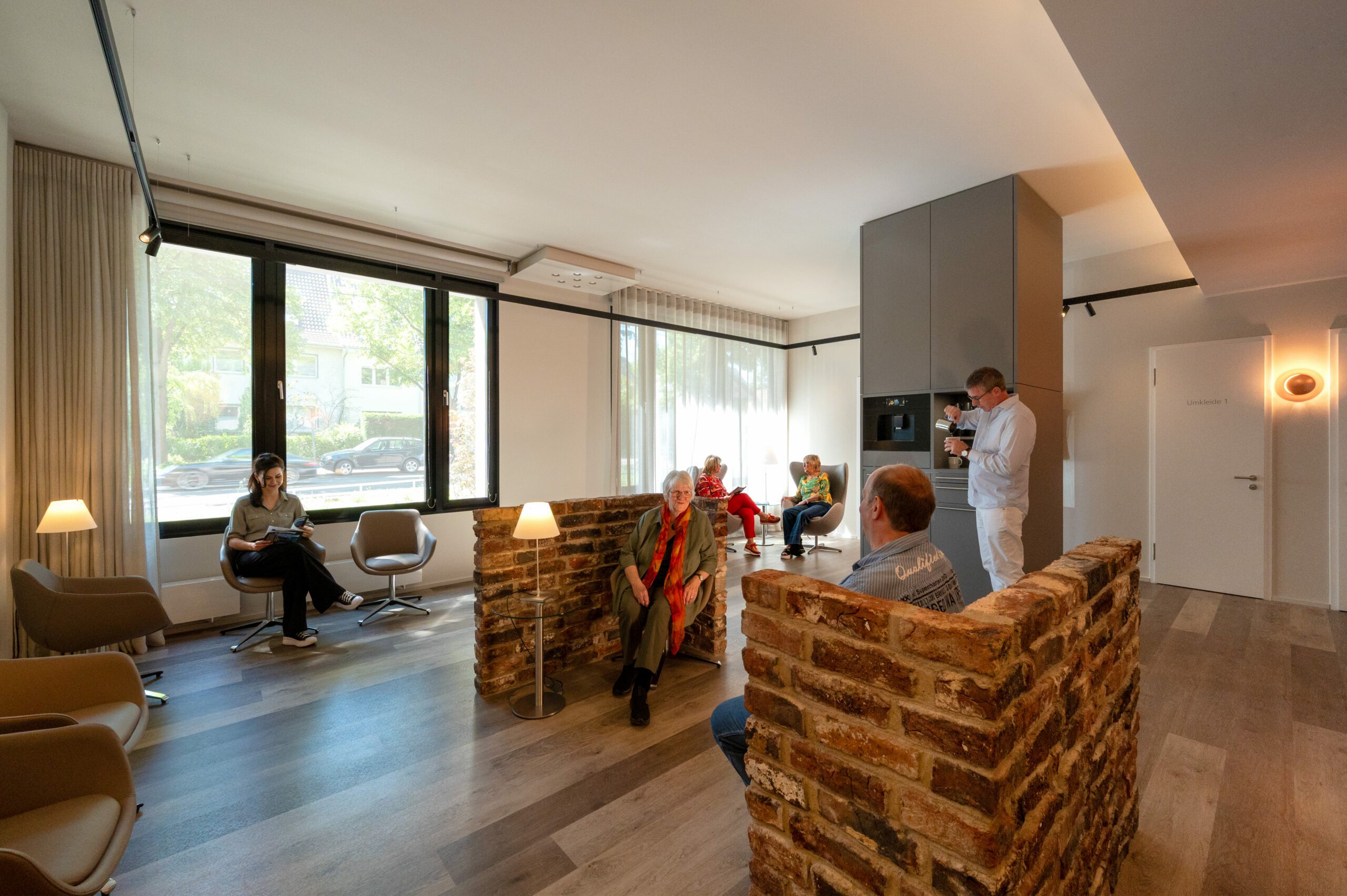Heller Wartebereich der mamonova Praxis in Lindenthal mit großen Fenstern, modernen Sitzbereichen und wohnlicher Atmosphäre. Mehrere Patient*innen sitzen entspannt, ein Mann bereitet sich einen Kaffee zu.
